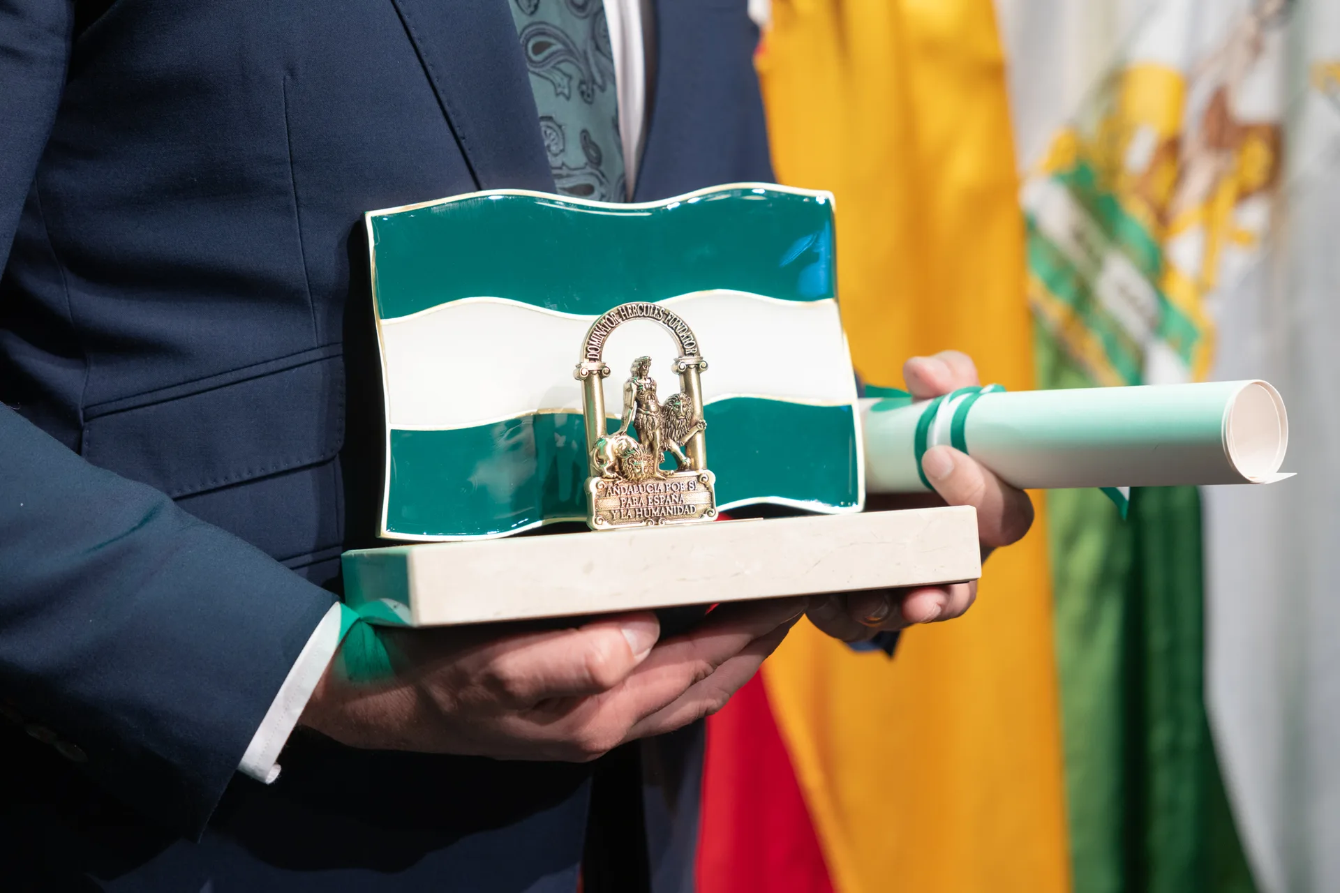 Las manos del director sostienen la Bandera de Andalucía de las Artes junto al diploma enrollado.