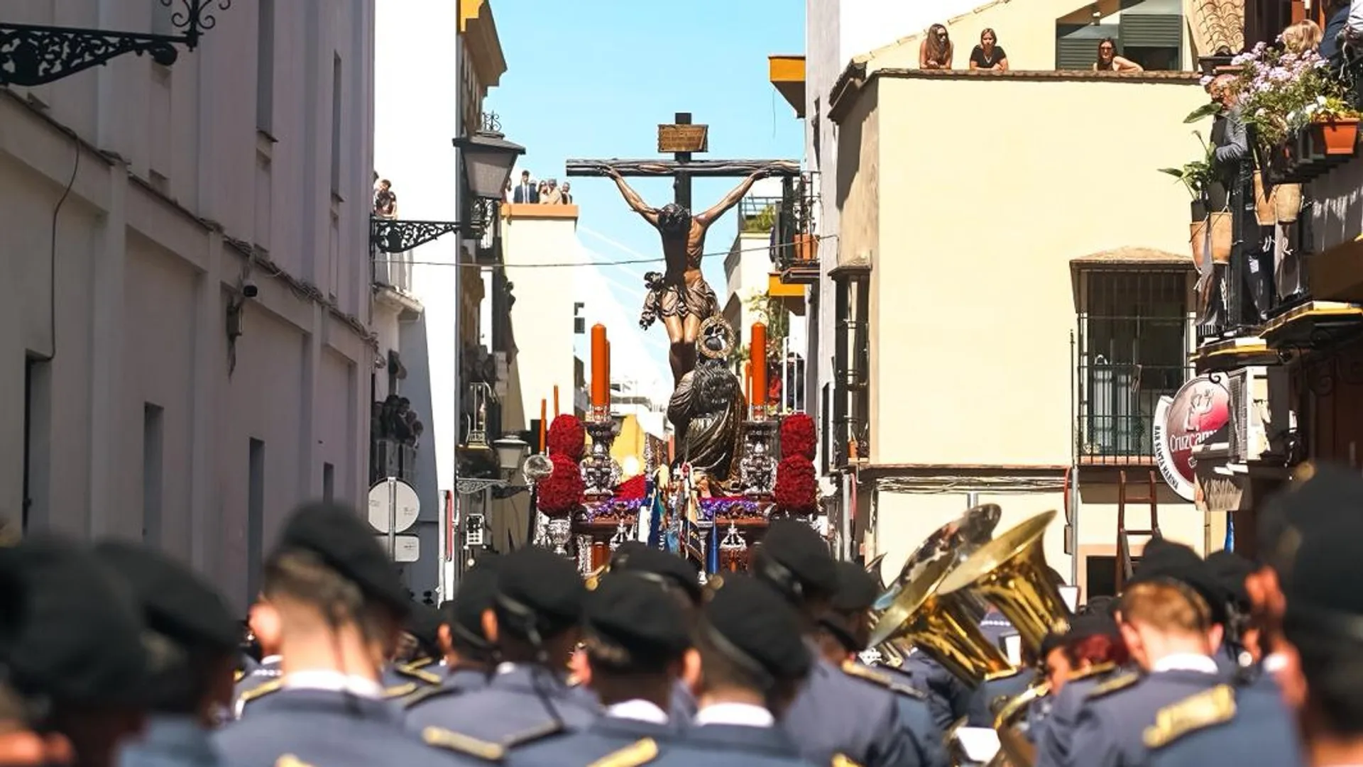 El Cristo de la Buena Muerte de la Hiniesta al fondo de una calle de Sevilla, con la banda de espaldas en primer plano.